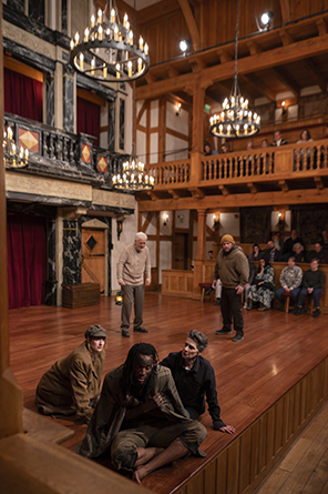 ASC_25_King Lear_014-Poor_Tom_et_al In deep vertical photograph from the corner of the Blackfriars Playhouse stage up to the upper balcony, chandeliers, and rafters, Edgar as Poor Tom sits crosslegged and barefoot with Lear's overcoat wrapped around him in the corner of the stage. Behind his left shoulder sits King Lear, behind his right shoulder sits the Fool. Gloucester stands in the background wearing a tan sweater and pants , and across from him stands Kent wearing his quilted jacket and stocking hat disguise. Behind them is members of the audience sitting on on-stage "Gallant stools" and in stage flanking seats at stage level and in the gallery above.